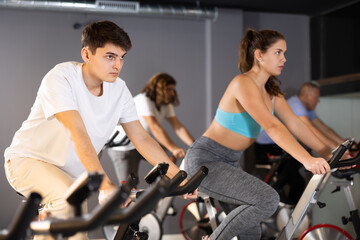 Fototapeta na wymiar Men and woman taking indoor cycling class at fitness center, doing cardio riding bike