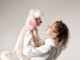 happy girl with a dog in a photo studio on gray. Curly woman in a white hoodie with a white poodle...