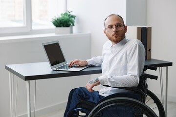 A man in a wheelchair looking at the camera businessman in the office working on a laptop online,...