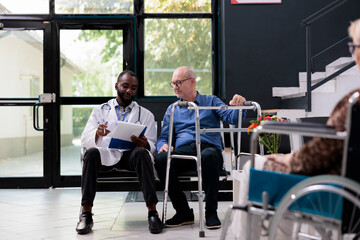 Practitioner medic holding clipboard with medical disease exam papers discussing health care treatment with disabled senior patient. People standing in hospital waiting area during checkup visit