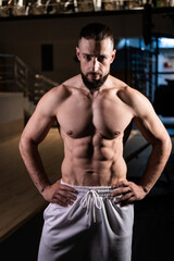 Fototapeta premium A handsome muscular male bodybuilder with a pumped-up torso with a beard and white pants looks at the camera