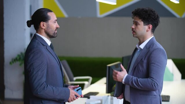 Two man employees in suits are talking in the office, a boss and a worker, solving work tasks. A man is holding a laptop.