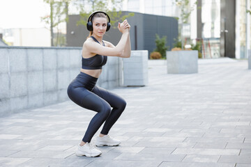 Determined athletic girl listening music, squatting during workout in city.
