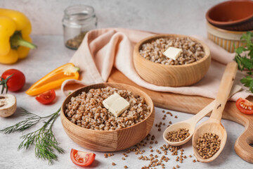 Wooden bowls of tasty buckwheat porridge with butter and vegetables on grey table