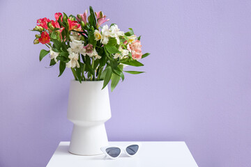 Vase with alstroemeria flowers and sunglasses on table near lilac wall