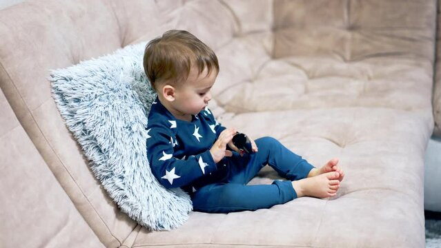 Lovely little toddler sitting cozy on the sofa. Nice kid in blue suit exploring the TV remote. Side view.