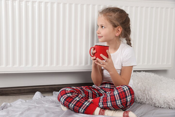 Little girl with cup of hot drink near heating radiator indoors, space for text