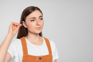 Young woman cleaning ear with cotton swab on light grey background. Space for text