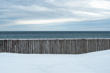 A wooden breakwater made of wood logs along the edge of the ocean with a bank of white snow in the foreground. The sky is cloudy with a blue tint. The blue ocean is in the background with a horizon   