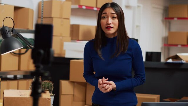 Young entrepreneur filming teleshopping video on phone, advertising products in warehouse space. Female worker using social media channel to create online ad promotion. Handheld shot.