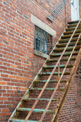 The exterior corner wall of an old brown and red colored brick building with rusty metal fire escape stairs. There's a multi-pane glass window in the building at the top of the vintage steps. 