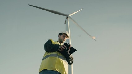 A professional Caucasian engineer in a reflective vest and white helmet uses a tablet to audit wind turbines in a field, symbolizing the growth of renewable energy. Eco-sustainability concept: energy.