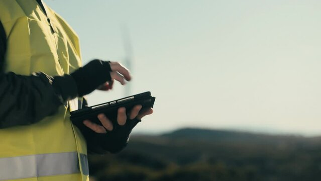 Promoting a greener future, an engineer wears a reflective vest while using technology software on a tablet to audit wind turbines in a field of renewable energy generators on a sunny day. Eco-theme.