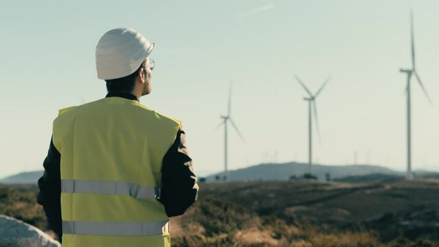 A white-helmeted engineer surveys wind turbines while walking, showcasing the technology behind renewable energy. Theme of integration of ecological principles with the development of renewable energy