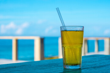 Big glass drink plastic straw green tea looks like yellow beer or kombucha blue sea white clouds background water drops condensed