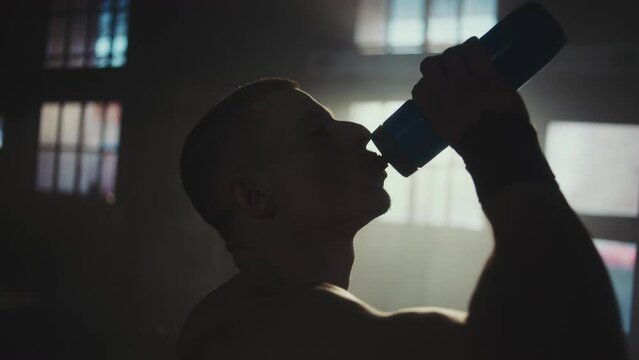 Topless muscular sportsman drinking water from a sports bottle in abandoned warehouse after doing some exercise in slow-motion. Tracking shot