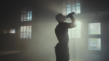 Slow-motion shot of a muscular young man standing in the gym after exercise and drinking water from plastic bottle - Powered by Adobe
