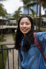 Young Asian girl taking a selfie or facetime video call on a smartphone outside in an urban area with palm trees