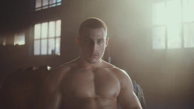 Young muscular man with six pack abs sitting on a large tyre looking at camera at gym
