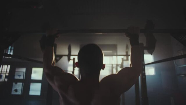 Slow-motion close-up view of a muscular young man pulls up exercising on bar at gym. Back view