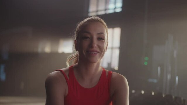 Young sportswoman with braces on teeth looking at camera and smiling while sitting on a rowing machine in slow-motion