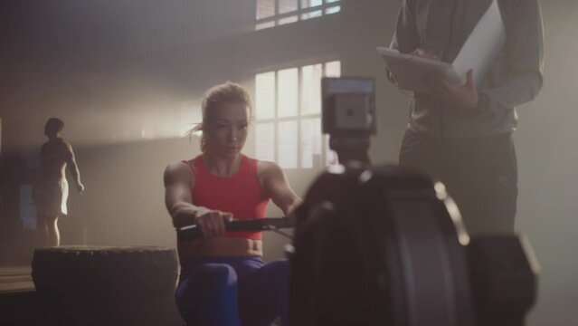 Extreme slow-motion shot of a beautiful woman working out on rowing machine with trainer. Muscular man skipping in the background