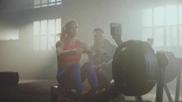 Male fitness trainer motivating female athlete to exercise on rowing machine at sports gym while a boxer shadow punching in the background