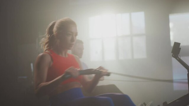 Attractive fitness couple training on rowing machine with great effort at sports gym. Muscular strong man working out with large tyre