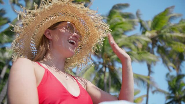 Beautiful elegant woman in straw hat waiving hand hello to someone, with scenic green palm trees on motion background. Girl in red swimsuit relaxing on beach smiling slow motion shot RED camera Hawaii