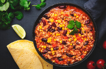 Chili con carne with beef, red beans, paprika, corn and hot peppers in tomato sauce, spicy tex-mex dish in a cooking pot, black table background, top view