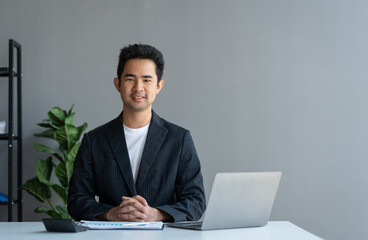 Young Asian businessman sitting with hands clasped on desk relaxing after Get financial information, statistics, graphs, income charts. while working online on laptop computer at office.