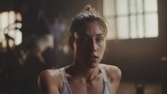 Slow-motion portrait of young beautiful sweaty woman laughing on camera in fitness centre. Close-up shot. People exercising in the background
