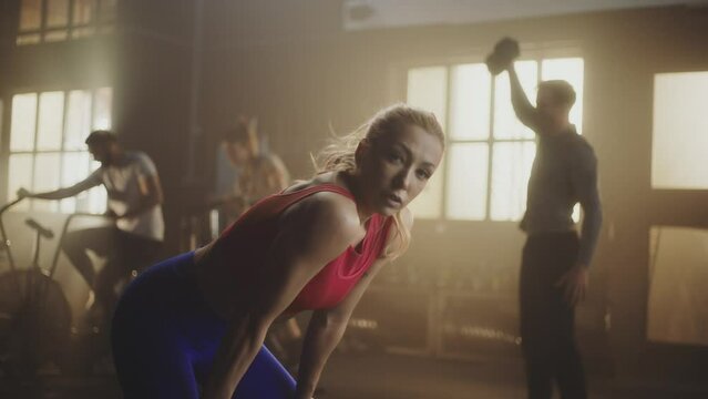 Strong woman resting after workout and looking at camera. Young sporty people are training together in background at gym
