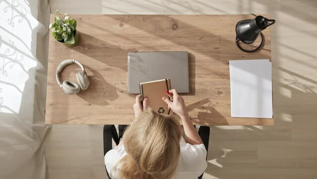 Top view, young blonde woman opens workbook and laptop while sitting at table and writes down first lesson. girl sits at home at her desk and computer, gets education.