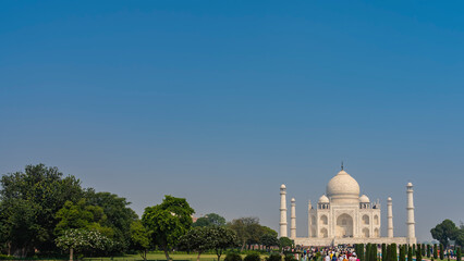 Obraz premium Beautiful white marble Taj Mahal against the blue sky. Green and flowering trees grow in the garden. A lot of people are walking along the paths to the mausoleum. Copy space. India. Agra