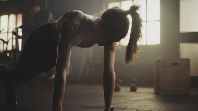 Slow-motion shot of a fit caucasian woman doing push-up exercise on the floor at gym. Two men working out in the background. - Powered by Adobe