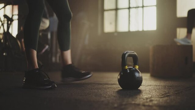Close-up leg view of a athletic woman lifting a kettelbell from floor at gym while another man cycling on exercise bike