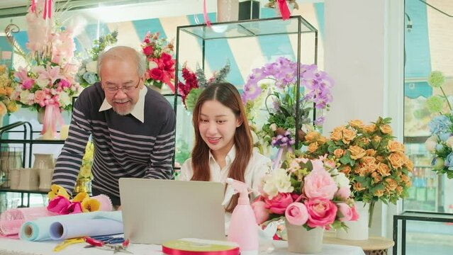 Asian young beautiful female florist worker discussing with an old man shop owner, website arrangement for online business service, happy work in colorful flower store, e-commerce SME entrepreneur.