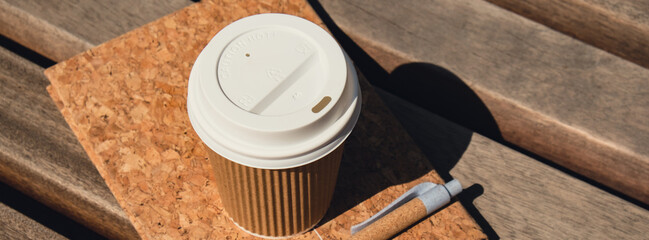 Eco recycling paper cup with coffee or tea on kraft paper with empty paper notebook on wooden bench. Concept of study work outdoors. Take away coffee to go. Copy space for text. Disposable Cardboard