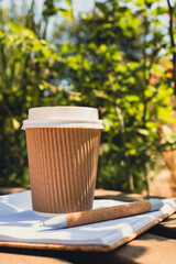 Eco recycling paper cup with coffee or tea on kraft paper with empty paper notebook on wooden bench. Concept of study work outdoors. Take away coffee to go. Copy space for text. Disposable Cardboard