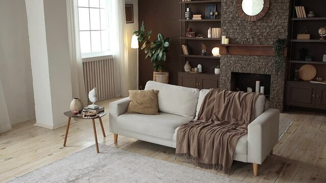 Modern stylish spacious living room with beige sofa on the background of large windows, brown wall, fireplace with shelving filled with books, decor and potted plants. Cozy chalet interior. Nobody