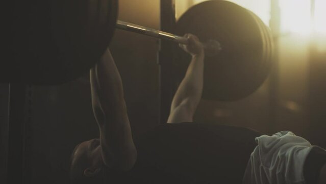 Handheld shot of a strong man lying on gym bench and lifting up barbell in slow motion at gym