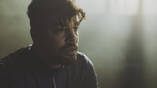 Close-up shot of a depressed young adult man sitting on a tyre and looking at something in gym. Slow-motion shot