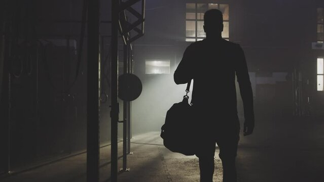 Slow-motion back view of a handsome guy with bagpack walking in an abandoned warehouse to exercise