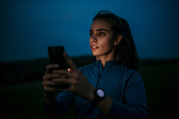 Pensive female athlete night runner standing in the dark and using the phone outside. Night jog concept