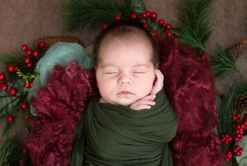Sleeping newborn infant wrapped in green surrounded by christmas holiday colors and greenery - 574160227
