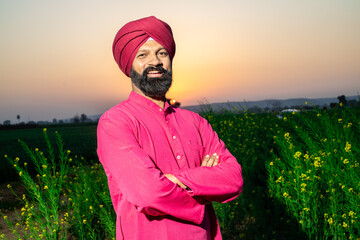 Portrait of happy young punjabi sikh man farmer standing cross arms wearing red turban and kurta looking at camera at agriculture field. - obrazy, fototapety, plakaty