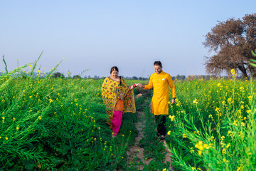 Fototapeta premium Portrait of Happy young indian punjabi couple wearing colorful outfit holding each others hand walking together in agriculture field.