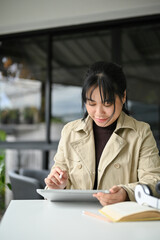 Asian female freelancer using digital tablet, focusing on her tasks, remote working at the cafe
