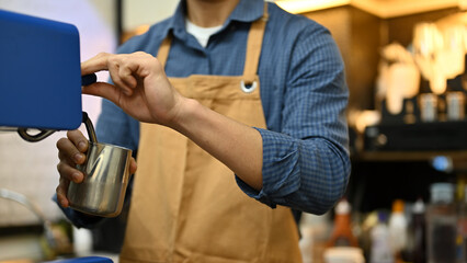 A male barista in apron working in the coffee shop, steaming fresh milk with machine